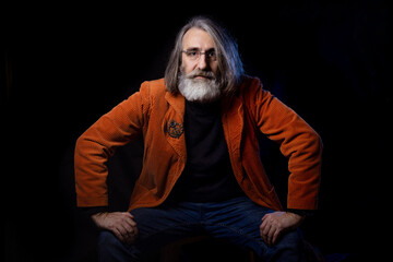 Portrait of a mature man in a stylish orange jacket on a black background . A fashionable intellectual looks into the camera with mockpice and interest. 