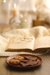 Open book, reading glasses, soft blanket and plate with chocolate and chocolate chip cookies. Hygge at home. Selective focus.