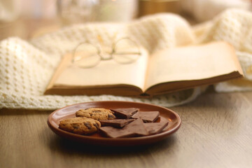 Open book, reading glasses, soft blanket and plate with chocolate and chocolate chip cookies. Hygge at home. Selective focus.