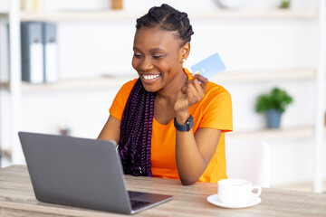 Online sales, e-commerce. Cheery black woman sitting at table with laptop and credit card, shopping...