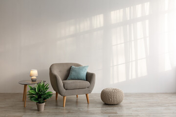 Comfortable armchair with pillow, luminous lamp on table, potted plant and ottoman on floor on gray wall background