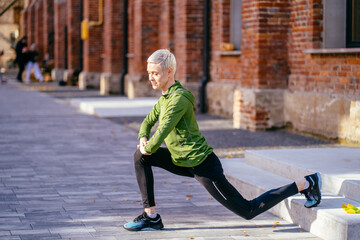 Middle age blond slim woman jogger in green activewear in an outdoor everyday sports routine. Female jogger workout, doing warming up exercise for legs.