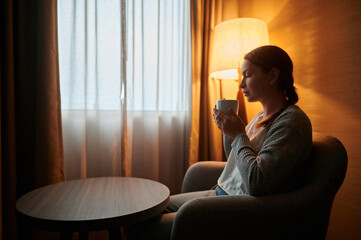 A young woman with a cup of tea sits in a chair near the window. The room has a soft light from a...