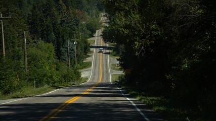 A white pick-up-truck driving in the middle of a lonely, rolling, american highway with double yellow lines. A power line is on the left side and trees are on either side.