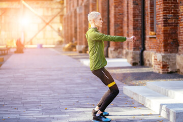 Preparation for running workout .Blond woman 40-45 years old training with fitness resistance band outdoor. Confident woman warming up enjoying workout in urban environment.