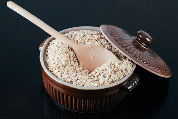 Oat flakes in ceramic pot with wooden spoon on dark table