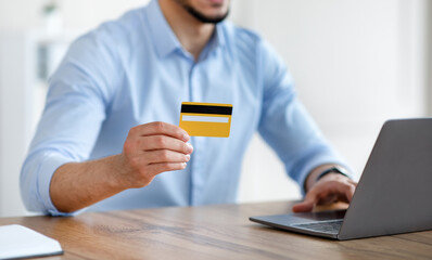 Unrecognizable Arab guy with laptop and credit card buying goods via internet at office, closeup