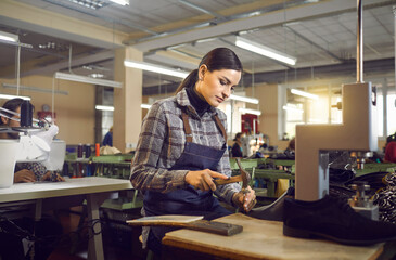 Shoe factory worker using professional tools to make new footwear. Young woman sitting at table in workshop and punching holes in leather material of future boots. Manufacturing industry concept