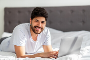 Always online. Young arab man working on laptop distantly, lying on bed and smiling to camera, free space