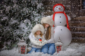 two sisters near the Christmas tree in warm hats and mittens and holding a bullfinch