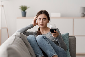 Sad indifferent caucasian young lady sits on sofa with glass of wine and suffering from headache in home