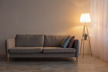 Cozy sofa, glowing lamp and plant in evening on gray wall background or living room, nobody