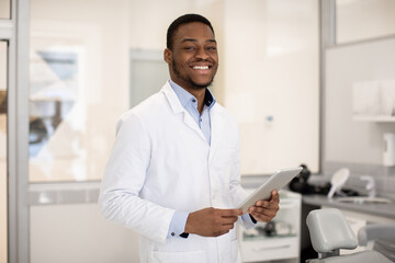 Dentistry Services. Handsome black male dentist with digital tablet posing in clinic