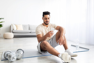 Handsome young Arab man with earphones and mobile device choosing music playlist for domestic sports training