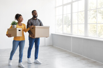 Fototapeta na wymiar Smiling young african american husband and wife carry cardboard boxes and plant, look at empty room and window