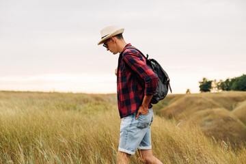 Naklejka na ściany i meble traveler with a backpack, walks in a field at sunset. Male tourist walks and enjoys the view of the beautiful nature