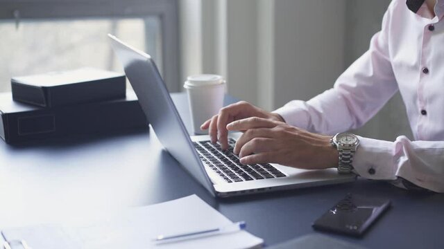 Young businessman working with laptop at modern office. Unrecognizable man typing on keyboard, writing email or message and drinking cofee at the table against grey concrete wall. Lateral motion shot