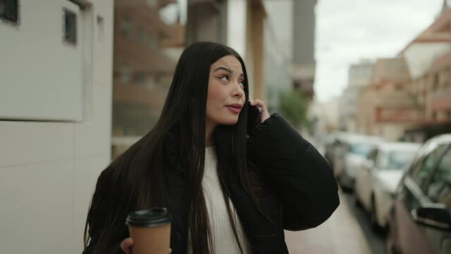 Young hispanic woman talking on the smartphone drinking coffee at street