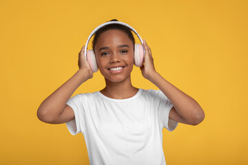 Happy teen pretty afro american girl in white t-shirt in headphones listening music, isolated on yellow background