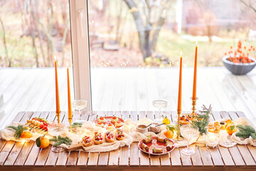 Christmas dinner feast. A small table is served with snacks, bruschettas, and canapes. A decorated dining table with champagne glasses, candles and christmas tree an garland in background
