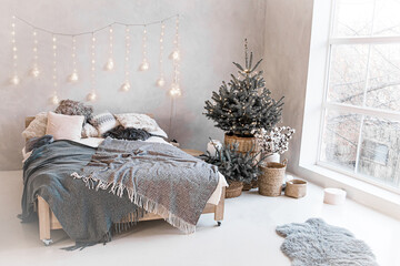 Bedroom with hugo-style bed with gray plaid. natural Christmas tree in pot with christmas lights.Festive decorated bedroom interior with window to floor ready for celebration of new year or Christmas