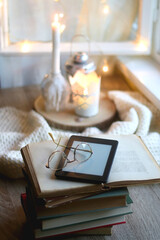 Stack of vintage boooks, digital e-reader, reading glasses, cozy blanket and lit candles. Hygge at home. Selective focus. 
