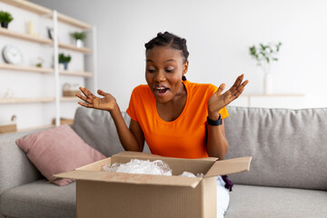 Excited black lady receiving parcel, opening cardboard box at home, satisfied with cool purchase