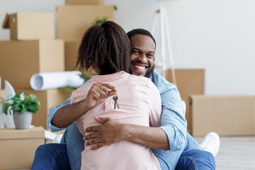 Obraz na płótnie Canvas Glad young african american male hugs female and shows keys in living room with cardboard boxes