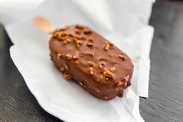 Macro closeup of one chocolate almond nuts ice cream bar on wood stick with plastic white wrap packaging package on kitchen table - obrazy, fototapety, plakaty