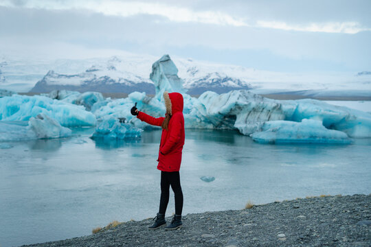 Girl traveler in red jacket with a hood  calling by video internet on smartphone leisure near Jokulsarlon glacial lagoon, Iceland. Female tourist takes photo selfie on cellphone on background.