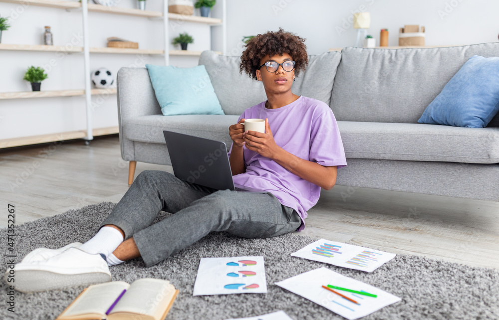 Wall mural thoughtful black teenager sitting on floor with laptop and study materials, taking break, drinking c