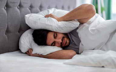 Upset arab man closing his ears with pillow, hiding from too loud sound or noise early in morning, lying in bed