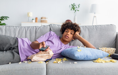 Lazy black teen guy lying on sofa with scattered food, holding TV controller, watching dull movie...