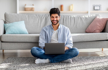 Arab millennial man using laptop computer, browsing internet, working online while sitting on floor...