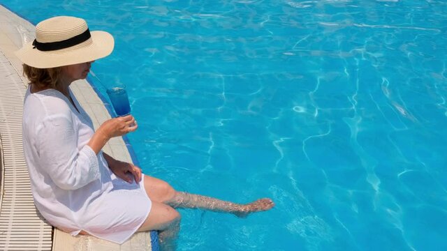 luxury senior woman dangling her legs in an outdoor swimming pool holding a blue refreshing cocktail wearing a straw hat. Summer rest. All inclusive