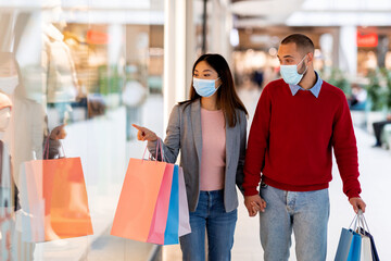 Young diverse couple in face masks shopping at supermarket, holding gift bags, pointing at shop window, selecting goods