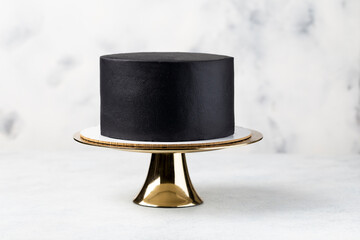 Black cake on golden cake stand, white table.