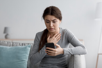 Scared depressed caucasian young lady sits on sofa look at smartphone and reading message at home