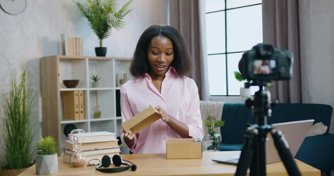 Adorable happy smiling surprised young african american woman opening gift box and taking out new smartphone while recording unboxing video for her internet vlog