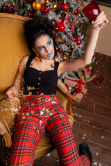 Fototapeta premium Brunette with art makeup in a dark top and checkered red pants with a Christmas tree in a photo studio