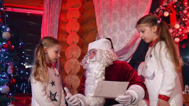 Children standing near Santa and talking to him. Kids sharing pictures and their Christmas dreams with Santa Claus. Santa asking girls about their wishes for holiday.