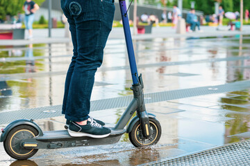 Close-up young person legs in jeans and in sneakers on electric scooter, wet asphalt with puddles...