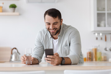 Smiling mature caucasian guy with beard drinking water and reading message on smartphone in scandinavian kitchen