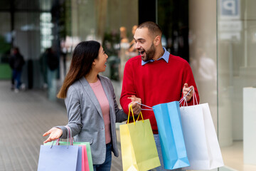 Excited international couple with gift bags standing near shopping mall, shouting OMG, overjoyed about seasonal sales