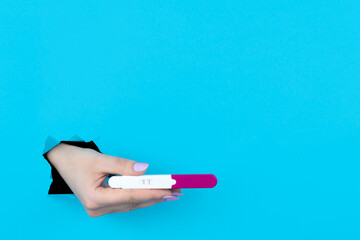 Female hand is holding pregnant test in torn hole of blue background. Medical healthcare gynecological, pregnancy fertility maternity people concept. Copy space for advertisement text inscription.