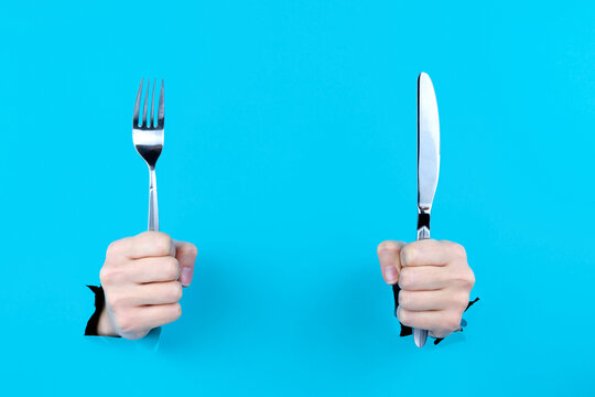 Female hands are holding cutlery metal knife and fork in torn hole of blue background. Food and restaurant menu concept. Serving dishes, lunch, waiting for order. Copy space for text inscription.