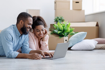 Smiling young african american woman and man looking at laptop with new design for room, lying on...