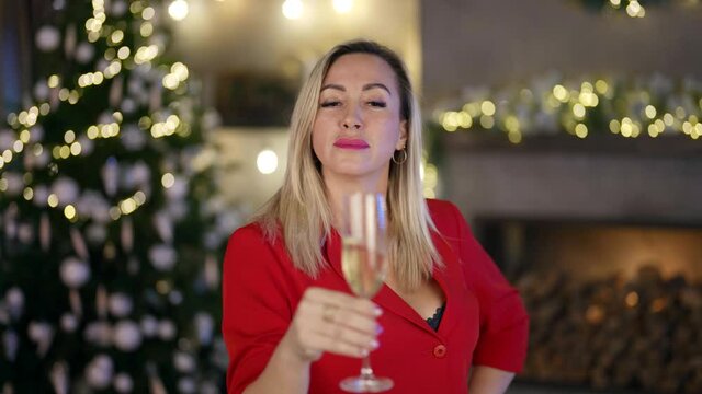 a blonde in a red blazer shakes wine in a glass and sips on the background of a decorated living room for Christmas