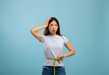 Sad and disappointed asian lady measuring waist with tape, unhappy about her body parameters, blue background