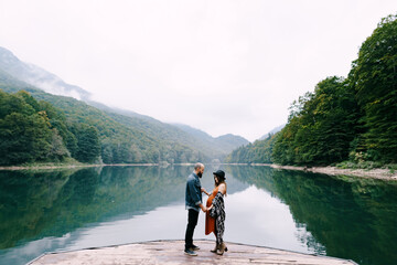 Pregnant woman and man stand holding hands on the pier by the lake